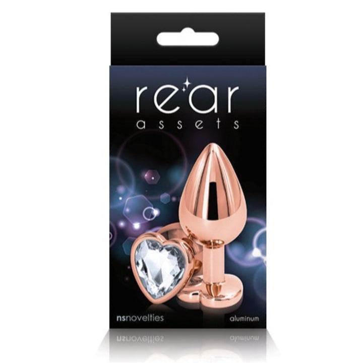 Image of the packaging for the Rainbow Heart Jeweled Metal Butt Plug in Rose Gold color with clear jewel. Text reads rear assets, NS Novelties, aluminum.