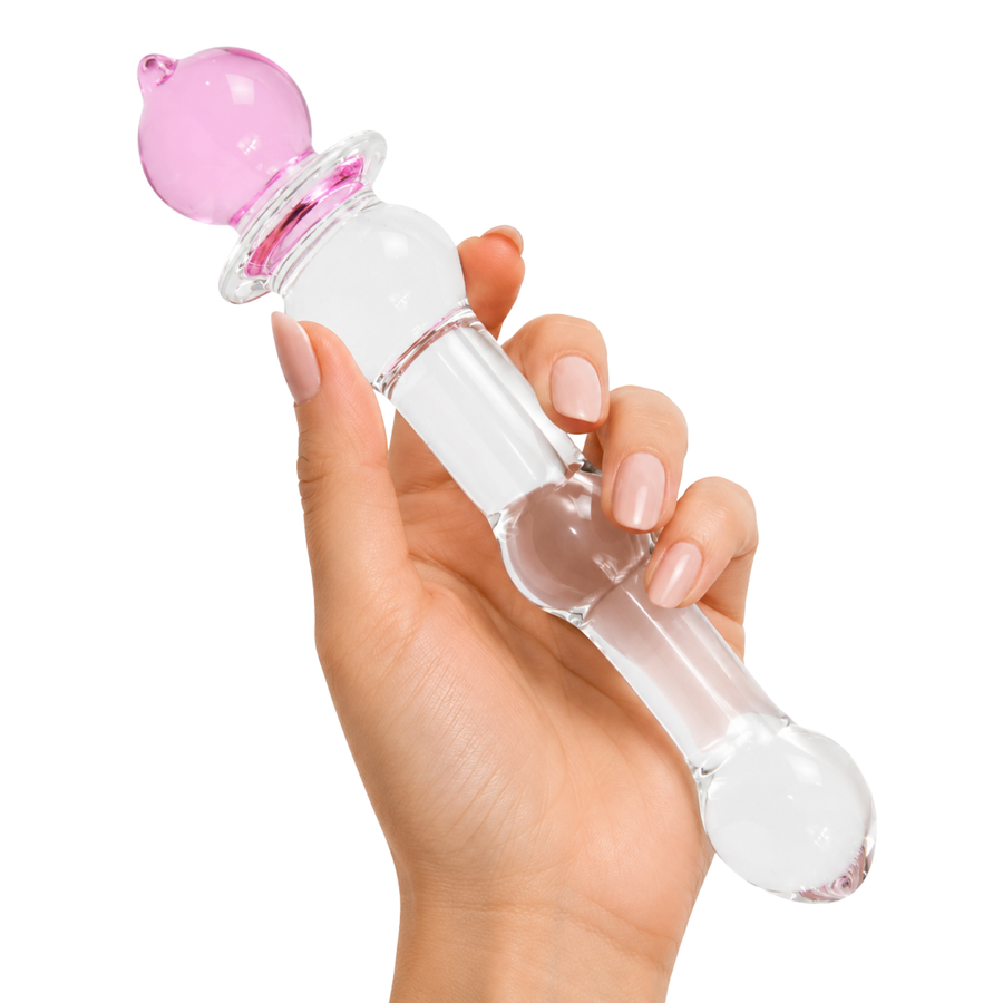 A hand with light pink manicured nails holds a clear glass dual-stimulation massager with a pink bulbous tip and a transparent cylindrical shaft on a solid white background.