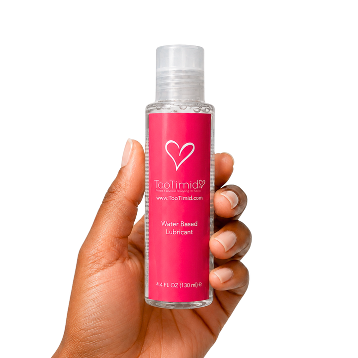 A hand holding a clear bottle of TooTimid 4.4 oz water-based lubricant with a bright pink label and a clear cap, set against a plain white background.