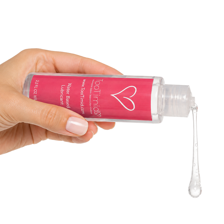 A hand with light pink manicured nails holds a clear plastic bottle of water-based lube against a white background, with a clear gel-like liquid dripping from the open cap.