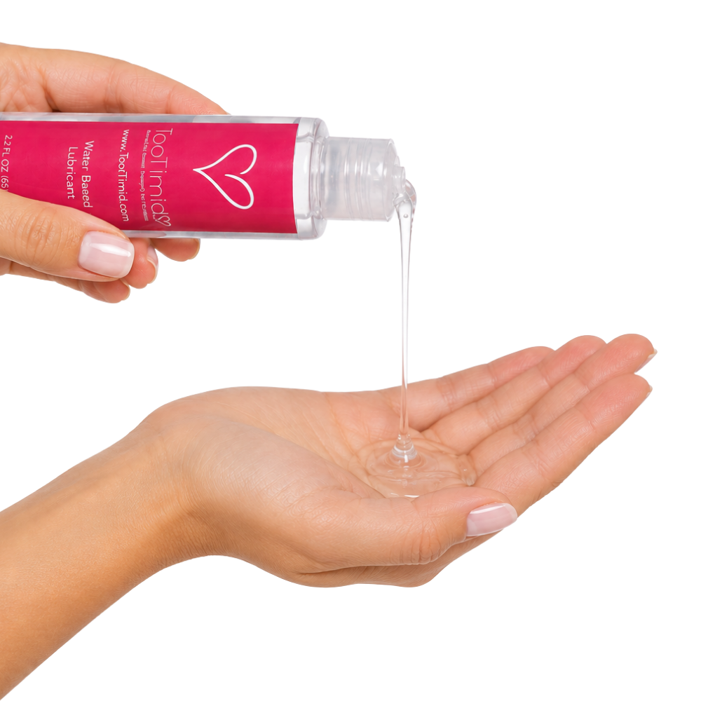 A hand holds a clear bottle with a bright pink label and white text, pouring a water-based lubricant into an open palm against a white background.
