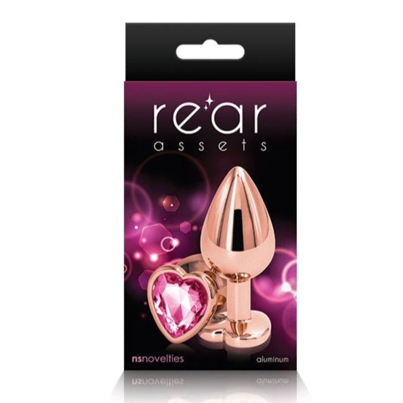 Image of the packaging for the Rainbow Heart Jeweled Metal Butt Plug in Rose Gold color with pink jewel. Text reads rear assets, NS Novelties, aluminum.