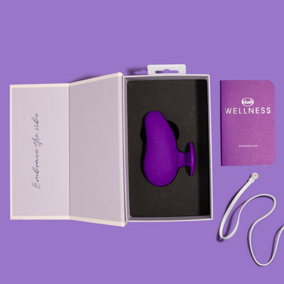 Blush Novelties palm vibrator in the white box open with black foam and white charging cable with instruction manual shown on purple background