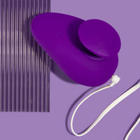 Blush Novelties palm adult sex toy vibrator with its white charging cord plugged in on a purple background 